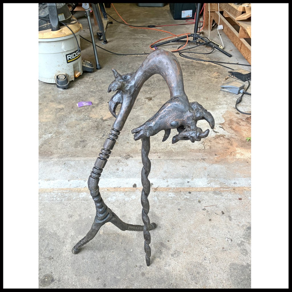 Rustic wood sculpture of dark, bent unicorn, facing downwards with spiraled main horn askew and acting as support. www.SawdustRiver.com