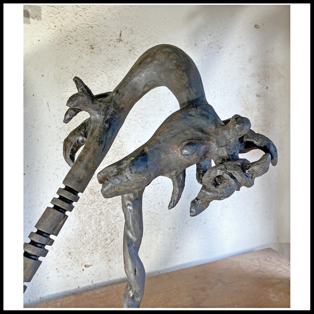 Rustic wood sculpture of dark, bent unicorn, facing downwards with spiraled main horn askew and acting as support. www.SawdustRiver.com